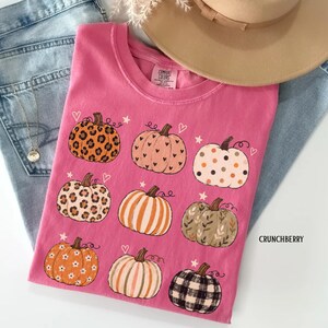 May include: A bright pink t-shirt featuring a graphic print of nine pumpkins with various patterns including leopard print, polka dots, stripes, floral, and plaid, interspersed with small stars and hearts. The text CRUNCHBERRY is printed on the side.