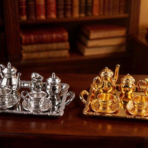 May include: Two miniature tea sets, one silver and one gold, are displayed on a dark wooden surface. Each set includes a teapot, creamer, sugar bowl, and cups with saucers, all arranged on a decorative tray. The background features bookshelves.