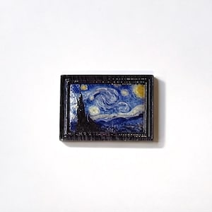 May include: A miniature framed artwork depicting Van Gogh's "Starry Night." The painting features swirling blue and white brushstrokes, a dark silhouette of a cypress tree, and a yellow crescent moon. The frame is dark brown.