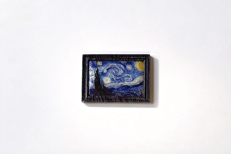 May include: A miniature framed artwork depicting Van Gogh's "Starry Night." The painting features swirling blue and white brushstrokes, a dark silhouette of a cypress tree, and a yellow crescent moon. The frame is dark brown.