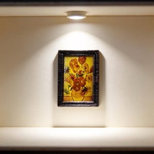 May include: Miniature framed painting of flowers in a vase, displayed in a small, illuminated box. The artwork features a vibrant bouquet with orange and yellow blooms against a yellow background. The frame is dark, and the box is a neutral color.