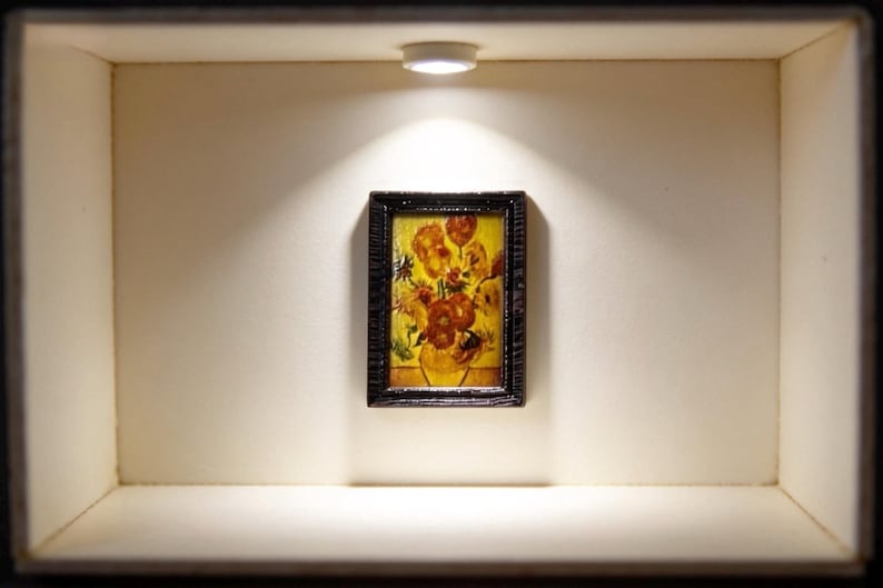 May include: Miniature framed painting of flowers in a vase, displayed in a small, illuminated box. The artwork features a vibrant bouquet with orange and yellow blooms against a yellow background. The frame is dark, and the box is a neutral color.