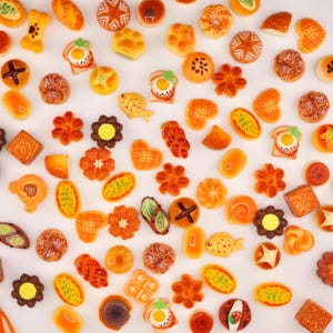 May include: A collection of miniature food items, including bread, pastries, and other baked goods. The assortment features various shapes and colors, such as orange, yellow, and brown. Some items have intricate details, like a fried egg on toast. The arrangement is on a white surface.