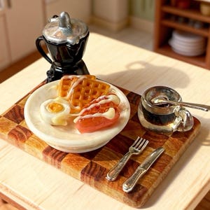 May include: Miniature breakfast scene featuring a plate with waffles, egg, and sausage, alongside a small coffee pot and a cup of coffee. A fork and knife rest on a wooden cutting board. The scene is set on a light-colored table.