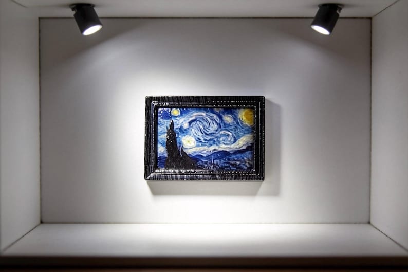 May include: Miniature framed artwork depicting Van Gogh's "Starry Night" painting. The artwork is displayed in a small, white, illuminated display case. The painting features swirling blue and yellow colors, with a dark, textured frame.