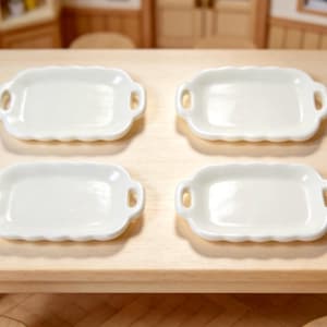 May include: Four small, white, rectangular serving dishes with scalloped edges and handle cutouts are arranged on a light wood table. The dishes are empty and appear to be made of ceramic.