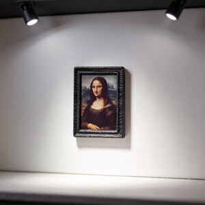 May include: Miniature framed print of the Mona Lisa, displayed on a white wall in a small gallery setting. The black frame surrounds the famous portrait. Two spotlights illuminate the artwork.