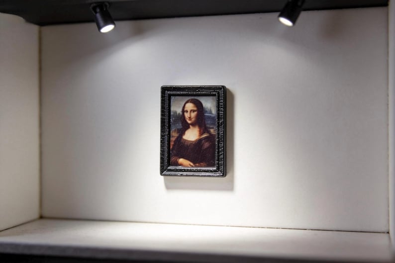 May include: Miniature framed print of the Mona Lisa, displayed on a white wall in a small gallery setting. The black frame surrounds the famous portrait. Two spotlights illuminate the artwork.