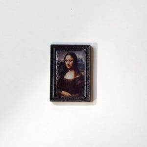 May include: A small, rectangular framed print of the Mona Lisa painting. The frame is dark brown with a textured appearance. The artwork features the iconic portrait of a woman with a subtle smile, rendered in warm, earthy tones.
