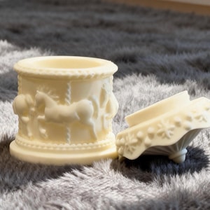 May include: Off-white carousel-themed trinket box with a matching lid. The box features embossed carousel horses and figures. The lid is decorated with floral patterns. The box is made of a solid, opaque material.