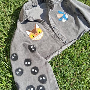 May include: Gray cropped denim jacket with button closures. The jacket features hand-painted cartoon character designs, including a cat face and small black soot sprites with white eyes. The jacket is lying on a bed of green grass.