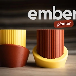 Könnte beinhalten: Zwei Keramik-Blumentöpfe, einer gelb und einer rot, mit einem gerippten Design. Die Töpfe sind mit "ember planter" beschriftet.