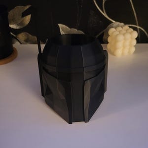 May include: A black plastic planter shaped like a Mandalorian helmet. The planter has a textured surface and a round opening at the top.