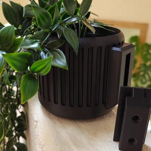 Puede incluir: Una maceta negra con un diseño acanalado y un soporte de pared negro. La maceta contiene una planta verde con hojas largas.