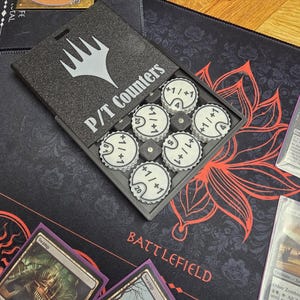 May include: A gray box with the text "P/T Counters" and a white logo, containing several white and black circular tokens. The tokens have numbers and the text "+1/+1" on them. The box is on a black playmat with the word "BATTLEFIELD".