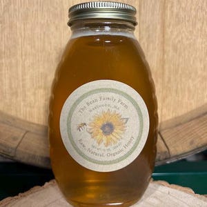May include: A jar of raw, natural, organic honey from The Bean Family Farm. The honey is a golden amber color and is in a glass jar with a silver lid. The label features a sunflower and a bee.