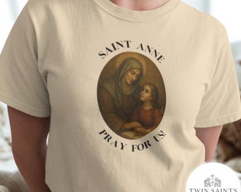 Op de afbeelding: Een beige t-shirt met een ovale afbeelding van Sint-Anna en een meisje, met de tekst "SAINT ANNE" erboven en "PRAY FOR US!" eronder. Het shirt heeft een ronde hals en korte mouwen.