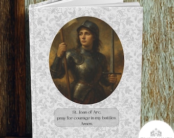 May include: A hardcover journal featuring an oval portrait of Joan of Arc in armor, holding a sword and staff. The cover has a floral pattern and text that reads "St. Joan of Arc, pray for courage in my battles. Amen."