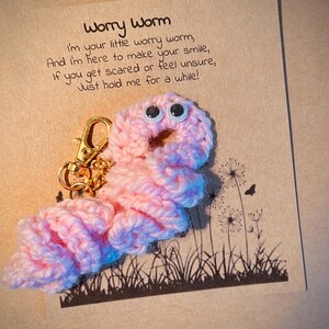 May include: Pink crocheted worry worm keychain with googly eyes and a gold chain. The keychain has a card attached with the text "Worry Worm I'm your little worry worm, And I'm here to make your smile, If you get scared or feel unsure, Just hold me for a while!"