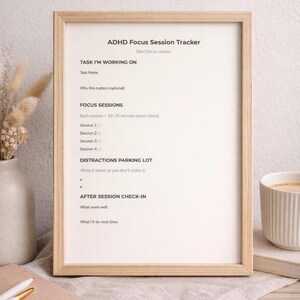 May include: A framed ADHD Focus Session Tracker with text including "Task I'm Working On" and "Focus Sessions." The tracker is in a light wood frame, with a neutral background. A cup of coffee and a vase are visible.