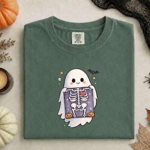 May include: A sage green crewneck t-shirt featuring a central graphic of a white ghost holding a purple X-ray showing a skeleton with a red heart. The design includes small orange pumpkins, a black bat, a skull, and yellow stars. The shirt has a Comfort Colors label.