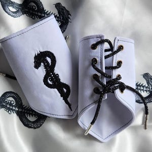 May include: Two white fabric wrist cuffs with black embroidered dragon designs. One cuff has a black lace-up closure with silver-toned aglets. The cuffs are displayed on a white satin fabric with a dragon pattern.