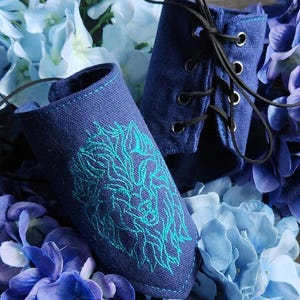 May include: Two navy blue fabric wrist cuffs with teal embroidery of a lion's head. One cuff is laced with black cord. The cuffs are surrounded by blue and purple hydrangea flowers.