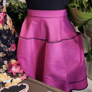 May include: A magenta-colored skirt with black stitching details. The skirt is displayed on a black mannequin and has a flared design. The word "MINT" is visible on a pink sign in the foreground. The skirt appears to be made of a smooth fabric.