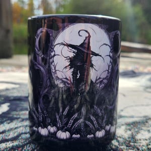 May include: A cylindrical black mug with a Halloween-themed design. The artwork features a scarecrow silhouette against a full moon, surrounded by trees, wheat, and pumpkins. The mug is likely ceramic and suitable for hot beverages.