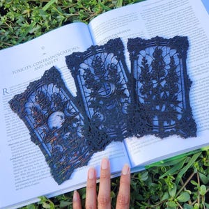May include: Three rectangular black laser-cut panels featuring intricate botanical and celestial designs, including floral motifs, crescent moons, and ornate filigree borders, arranged in a fan shape over an open book with visible text.