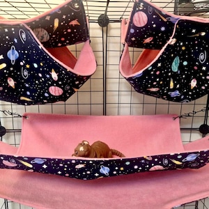 May include: Three pet hammocks and a bed inside a cage. The hammocks are black with a space-themed pattern of planets, stars, and rockets, and pink lining. A pink bed is below the hammocks, with a small brown toy animal resting on it.