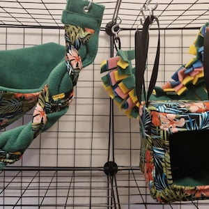 Puede incluir: Dos camas colgantes para mascotas con estampado de hojas tropicales. Las camas son verdes y presentan una variedad de colores, incluyendo rosa, amarillo y azul. Las camas están colgadas de una jaula de alambre negro.