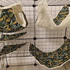 May include: A selection of pet hammocks and triangular beds. The items display a dark green fabric with a jungle pattern of leaves and cheetahs, complemented by a soft, cream-coloured lining. The accessories are designed to hang inside a cage.