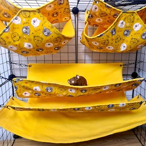 Puede incluir: Hamacas y ropa de cama amarillas para mascotas con un patrón repetido de caras de osos de peluche. Las hamacas están suspendidas de una jaula de alambre negra. Una pequeña mascota marrón descansa en una de las hamacas.
