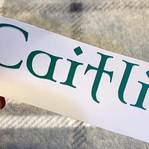 May include: A white sticker with the name "Caitlin" printed in a dark teal, elegant font. The sticker is held by a hand with a red painted fingernail and a gold ring.