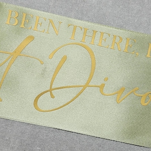 Könnte beinhalten: Olivgrüne Satinschärpe mit goldfarbenem Schriftzug. Der Text lautet „Been There. Done It. Got Divorced.“ Die Schärpe ist leicht angewinkelt, mit einem kleinen Zweig Grünzeug auf der linken Seite. Der Text ist in einer Schreibschrift.