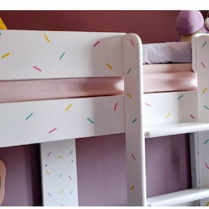 May include: White bunk bed with a pink and white patterned headboard and a white ladder. The headboard has a sprinkle design in yellow, pink, and blue.