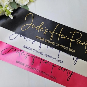 Może przedstawiać: Trzy satynowe szarfy w kolorze czarnym, białym i różowym z tekstem "Jude's Hen Party" i "Bride Squad Cyprus 2024" w złotych, czarnych i fioletowych literach.