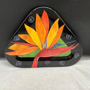 May include: A black, triangular decorative plate featuring a vibrant bird of paradise flower design. The flower has orange, yellow, and red petals with a green base. The plate is adorned with blue spiral accents.
