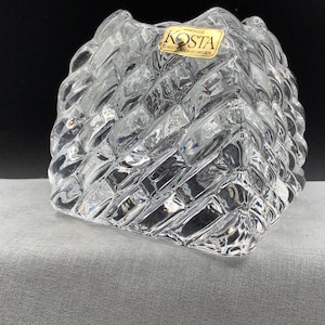 May include: Clear glass vase with a textured, diamond-patterned exterior. The vase has a square base and a slightly irregular top. A gold and white label reads "KOSTA HANDMADE KOSTA BODA AB SWEDEN".
