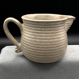 May include: A beige ceramic pitcher with a handle and a spout. The pitcher has horizontal, textured lines around its body. The handle curves upward and attaches to the body. The spout is on the opposite side of the handle.