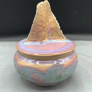 May include: A handcrafted ceramic jar with a lid. The jar is round with a lid that has a unique, textured, triangular handle. The jar features a blend of colors, including peach, purple, and green, with a glossy finish. The jar is suitable for storing small items.
