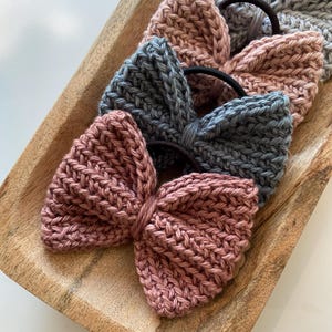 May include: Close-up of several knitted bow hair ties in shades of pink and gray. The bows are made of a textured knit fabric and are attached to black elastic bands. The hair ties are arranged in a wooden tray.