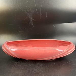 May include: A red-orange ceramic planter with an elongated, oval form. The planter features a raised, ridged pattern around the edge. The inside is smooth and open. Suitable for indoor or outdoor use.