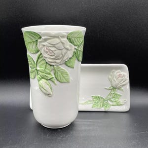 May include: White ceramic toothbrush holder and soap dish set, both adorned with raised rose and leaf designs. The roses have a delicate pink hue, and the leaves are green. The set is a bathroom accessory.