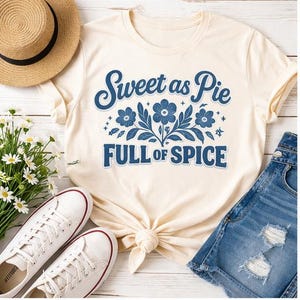 May include: Cream-colored t-shirt with the text "Sweet as Pie FULL OF SPICE" in blue. The design includes floral elements. The shirt is styled with denim shorts, white sneakers, and a straw hat.