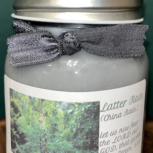 Op de afbeelding: Een grijze sojakaars in een glazen pot met een zilveren deksel en een grijze strik. Het etiket toont een boslandschap en de tekst "Latter Rain (China Rain)" en "16 oz. Soy Candles".