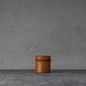 Peut inclure: Un petit récipient cylindrique avec un couvercle, fabriqué en bois brun clair. Le récipient est posé sur un fond gris, le couvercle étant légèrement plus foncé que le corps. Le design est simple et minimaliste, suggérant une attention aux matériaux naturels.