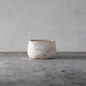 May include: A small, handcrafted ceramic bowl with a textured, off-white glaze. The bowl has a rounded square shape and a visible brown rim. The surface has a speckled appearance, and the bowl sits on a gray surface.