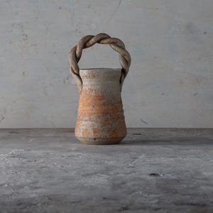 May include: A handcrafted ceramic vase with a twisted handle. The vase has a cylindrical shape with a textured surface and a warm, earthy color palette of beige and orange. The handle is a braided design.